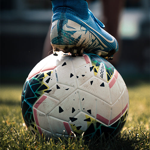A soccer cleat holding a soccer ball in place on a grassy field.